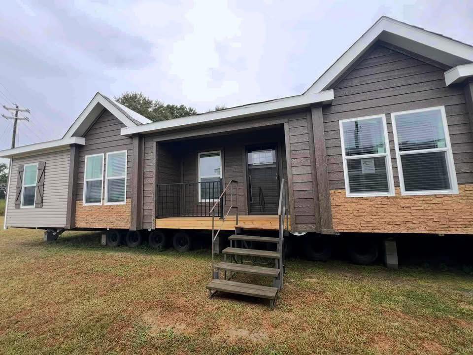 Modern manufactured home with brown siding, large windows, and a wooden porch. It sits on grass with a cloudy sky, conveying a cozy, rustic feel.