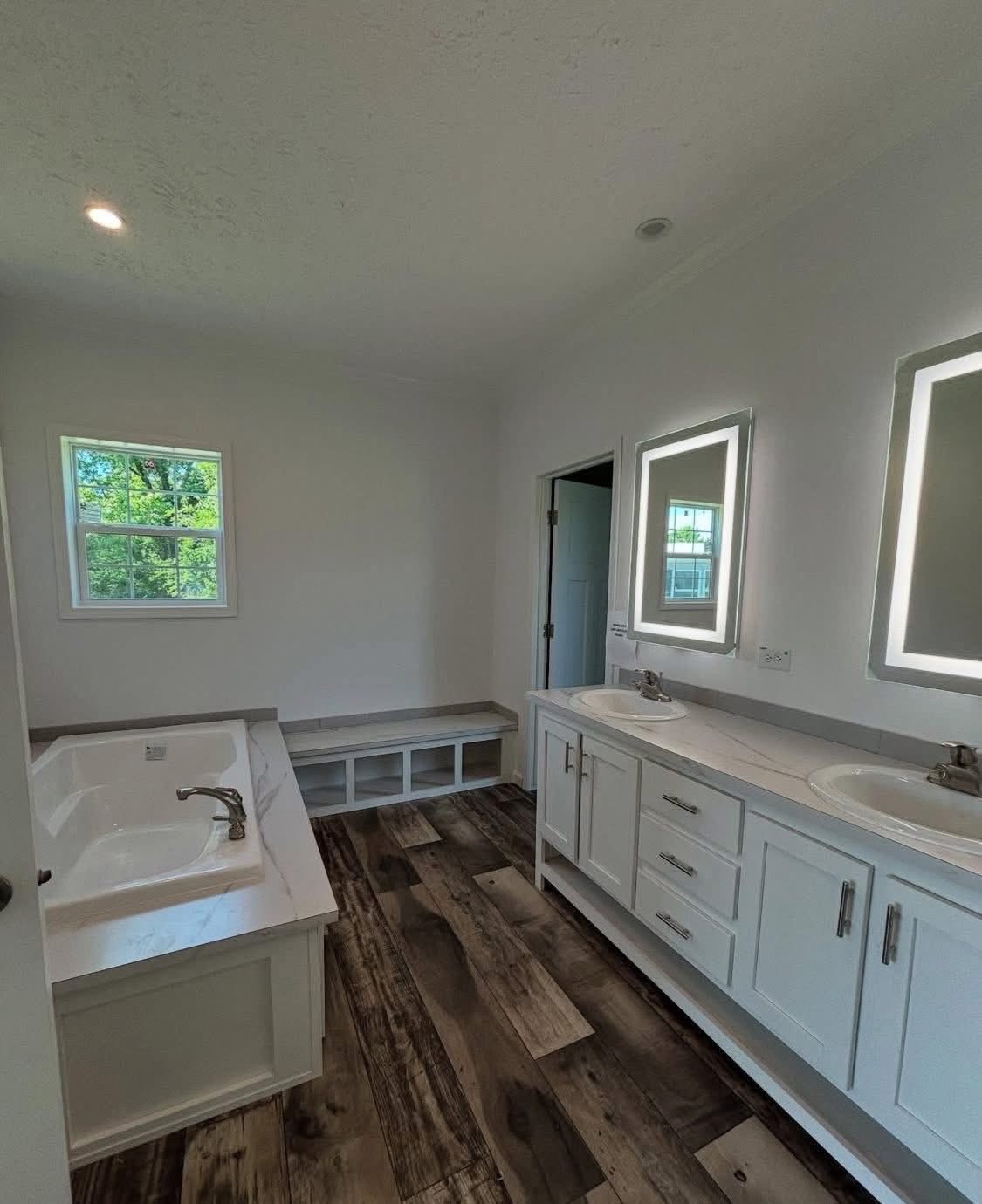 A spacious bathroom with wood-patterned tile flooring, a white double vanity with illuminated mirrors, a bathtub, and a small window. Bright and modern ambiance.