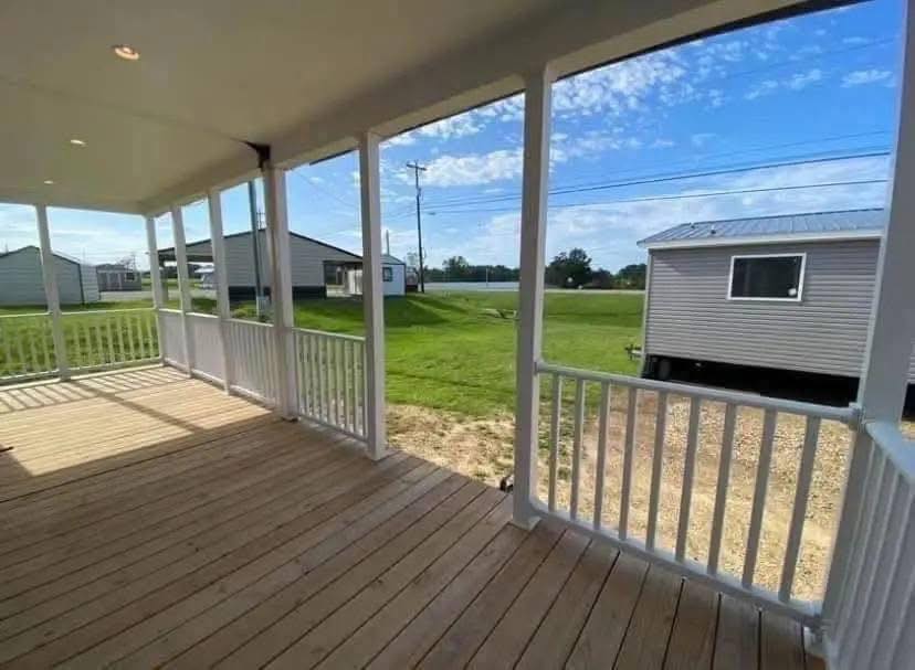 Covered wooden porch with white railings on a sunny day, overlooking green grass and small houses. The sky is clear with scattered clouds.