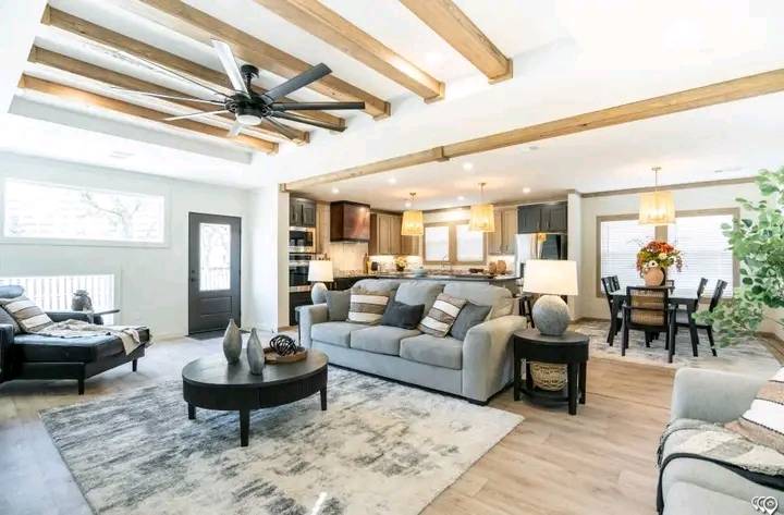 Spacious living room with cozy gray sofas, wooden beams, and a ceiling fan. Open kitchen and dining area with warm lighting and modern decor.