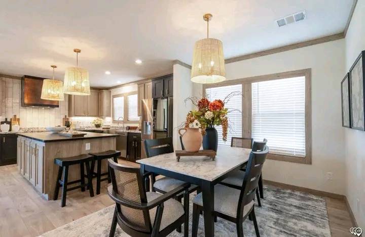 Modern kitchen and dining area with marble-topped tables, cushioned chairs, pendant lights, and a vase of vibrant flowers on a rug. Bright and inviting.