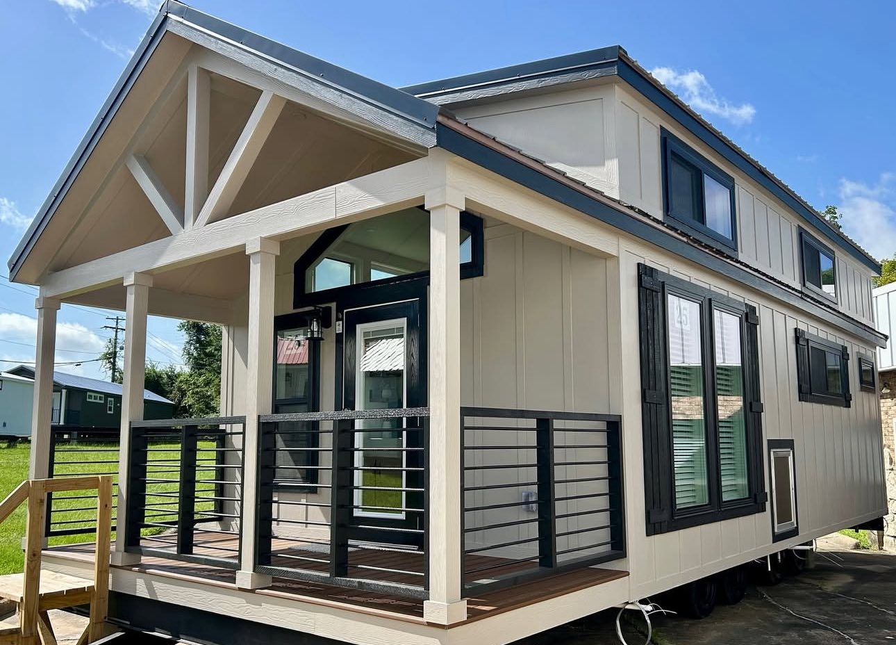 Modern tiny house with beige siding and dark trim, featuring a covered porch with a railing. Set against a bright blue sky, it conveys a quaint and cozy atmosphere.
