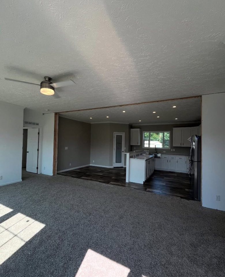 Open layout showing a spacious living room with carpet, ceiling fan, and an adjacent kitchen with white cabinets and a large window. Bright and airy.