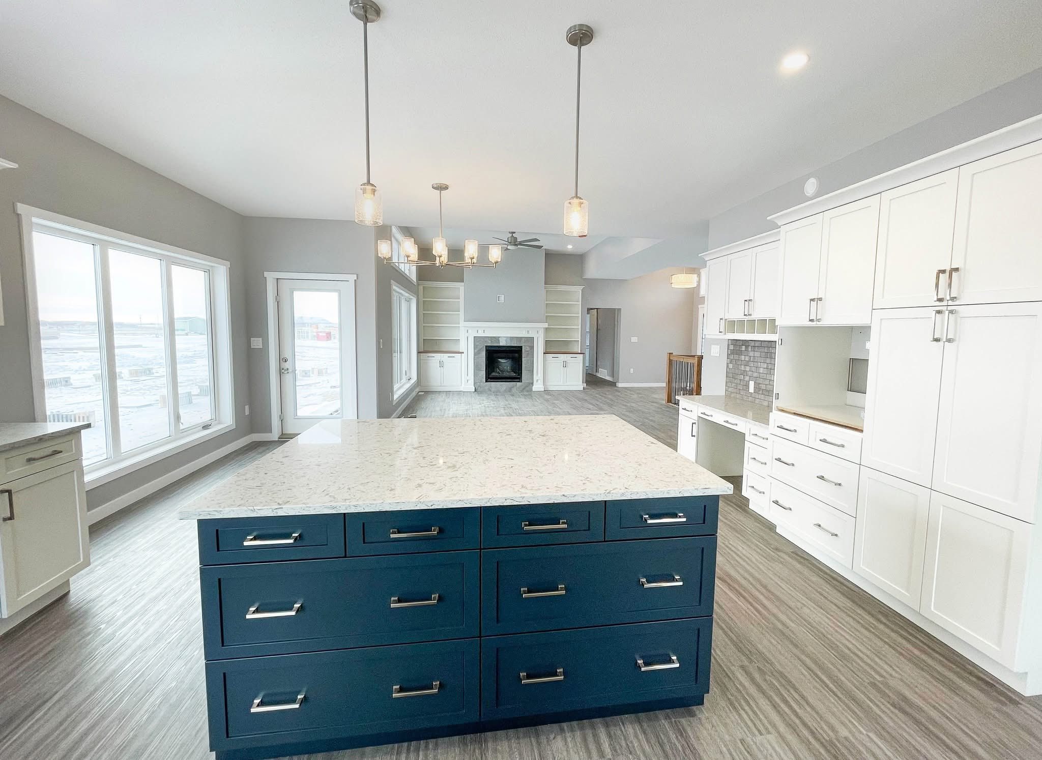 Spacious kitchen with blue island, white quartz countertop, white cabinets, pendant lights, large window, and open living area with fireplace.