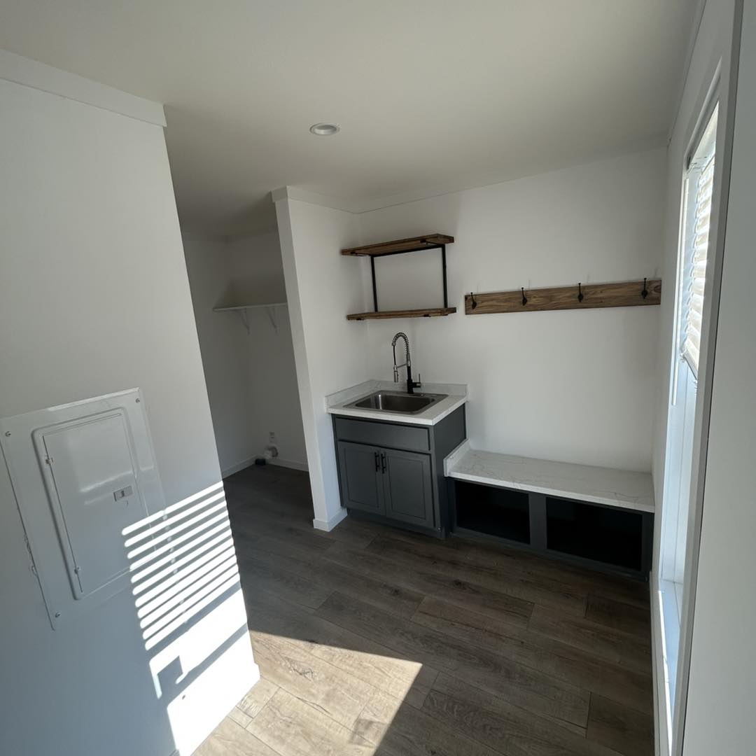 Compact room with wood floors, white walls, and natural light. Features a small sink with grey cabinets, wooden shelves, and hooks. Minimalist and clean.