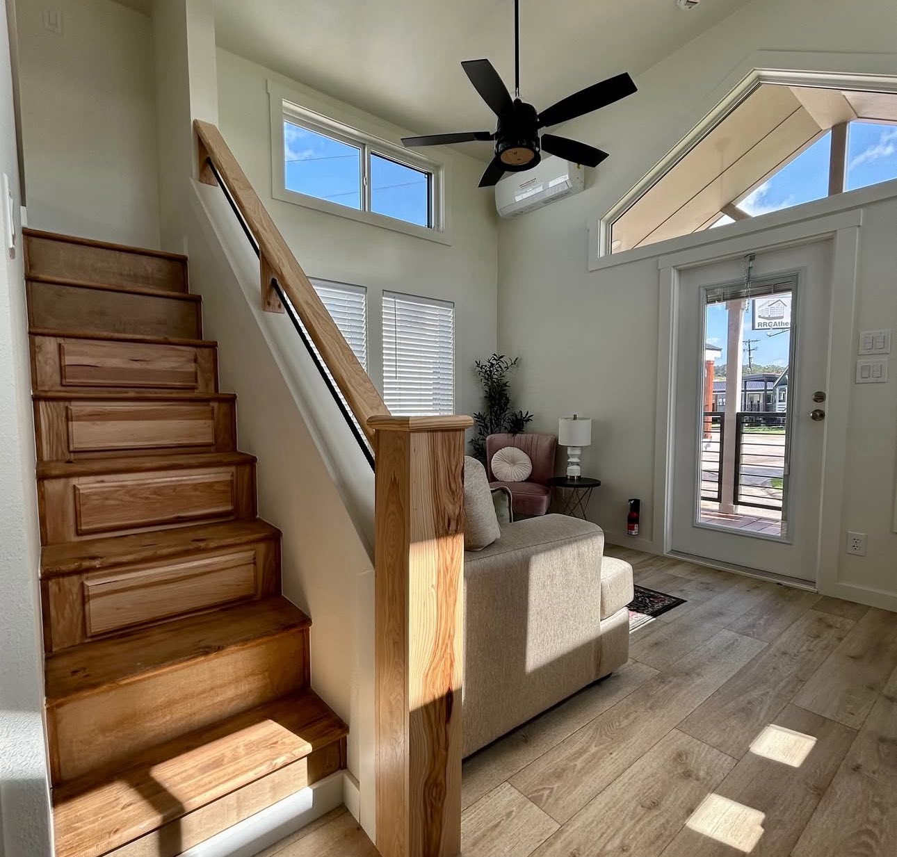 Sunny living room with wooden stairs, beige sofa, and black ceiling fan. Large windows and glass door illuminate the space, creating an airy feel.