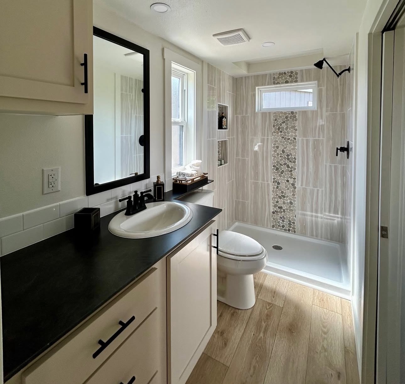 Modern bathroom with light wood flooring, a black countertop with white sink, and a shower featuring beige tiles and a pebble accent strip.