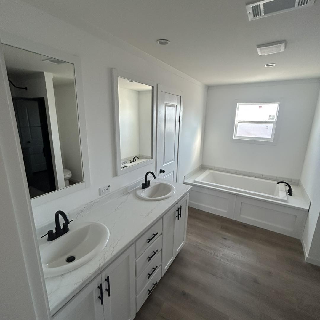 Bright, modern bathroom with double sinks on a marble countertop, black faucets, large mirrors, a spacious bathtub, wood floor, and white walls.