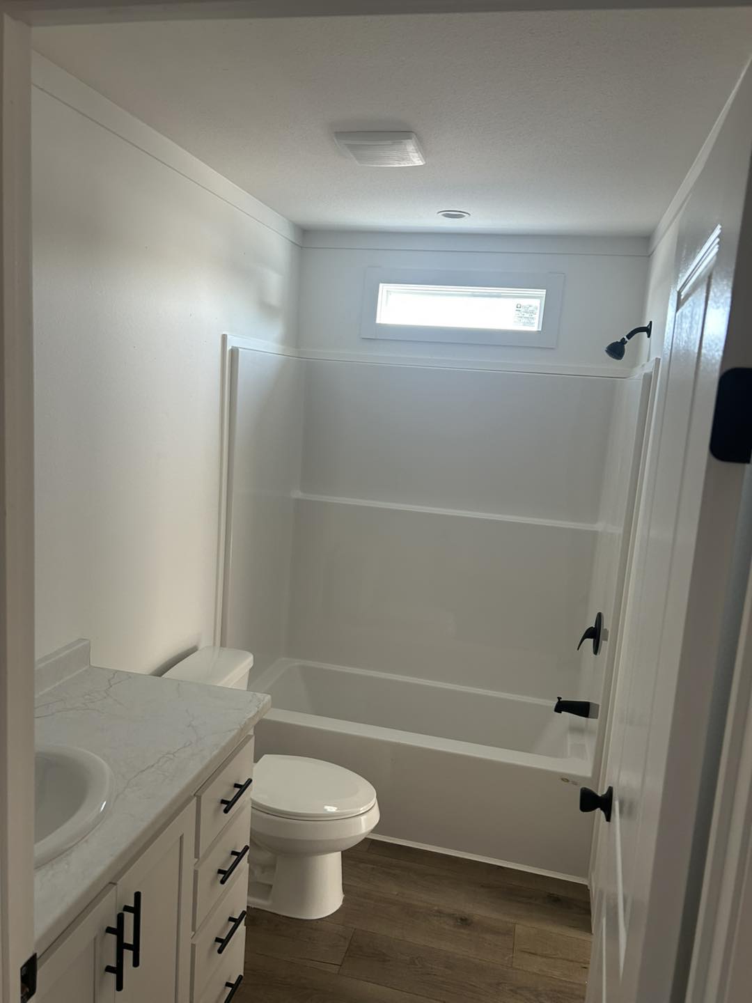 Modern bathroom with white walls, a bathtub-shower combo, small high window, wooden floor, marble countertop, and black fixtures, conveying a clean, minimalist style.