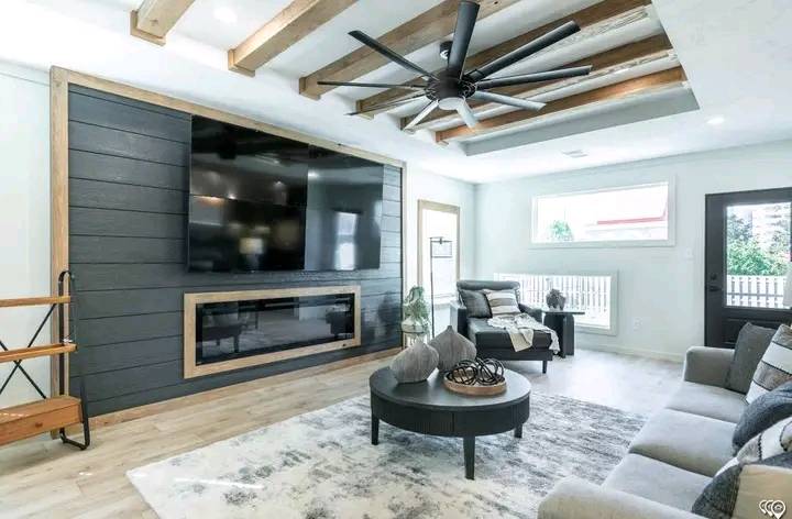 Modern living room with a large ceiling fan, black TV wall, and sleek furniture. Light tones, cozy vibe, with natural light from a window and door.