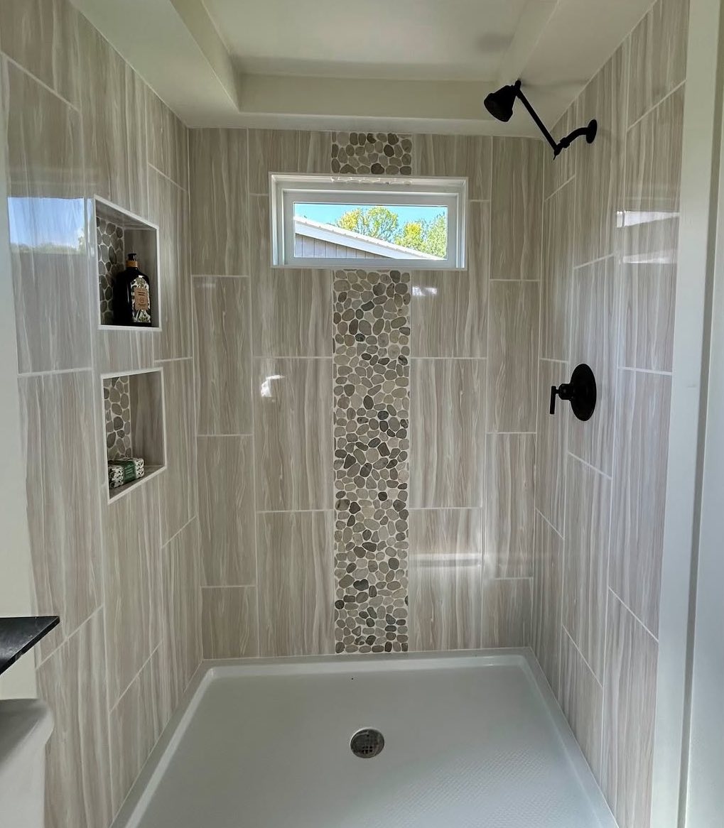 Modern shower with beige tiles, pebble mosaic accent, black fixtures, and built-in shelves. Natural light streams through a small window.