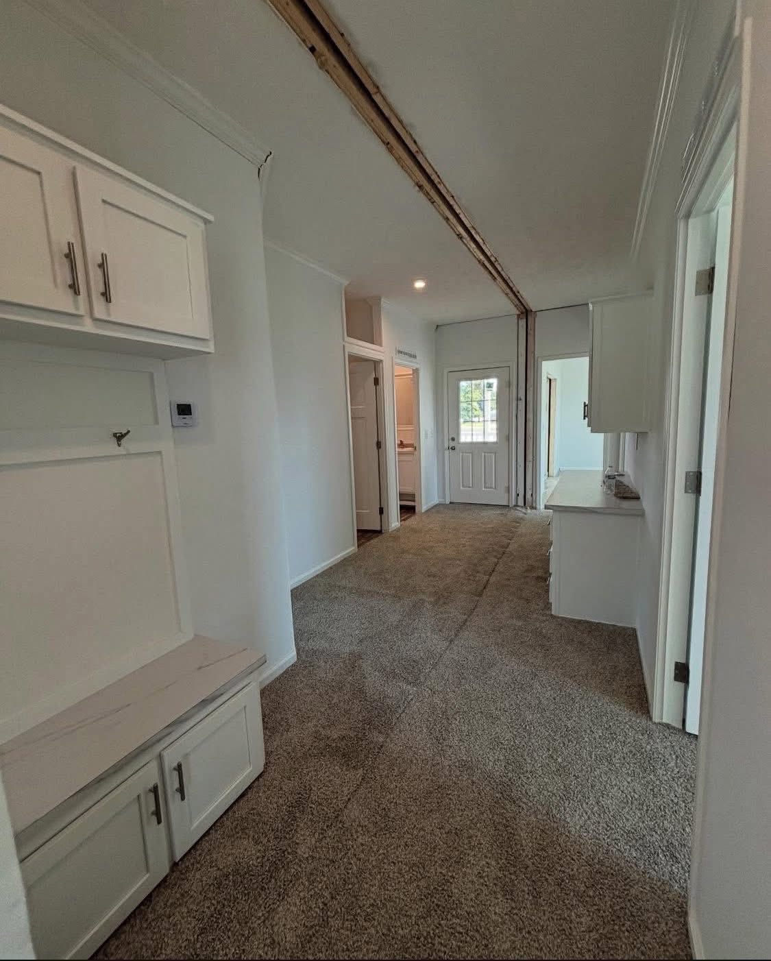 A narrow hallway with beige carpet, white walls, and ceiling. Cabinets with silver handles are on both sides. A door with a window is at the far end.