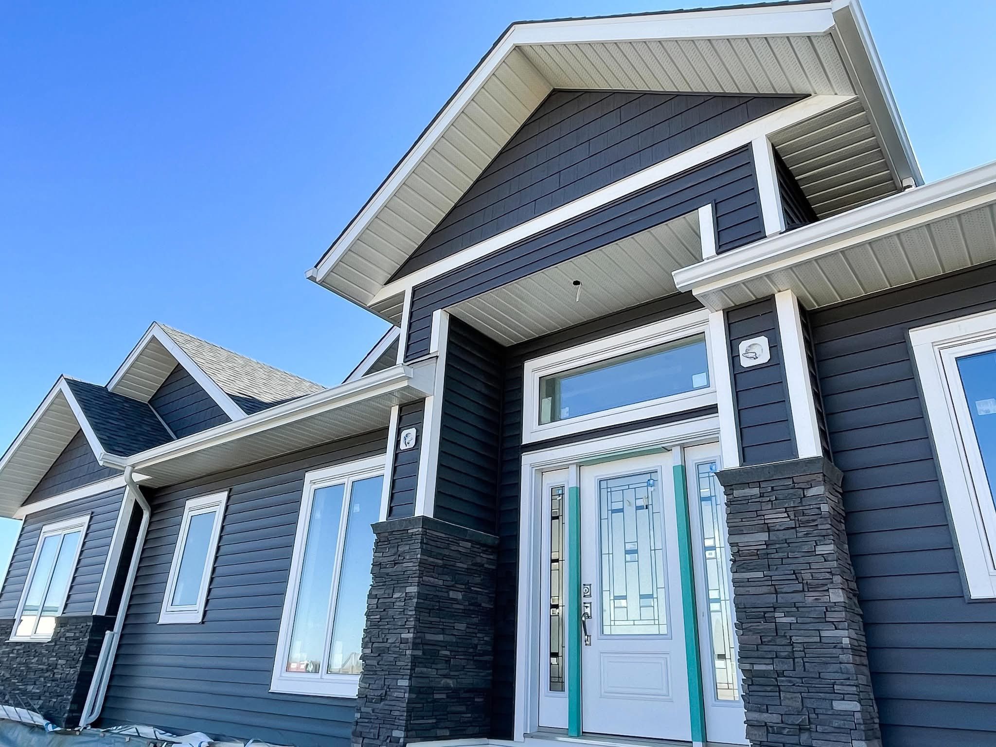Modern house exterior with dark gray siding and stone accents. Large windows and a glass-paneled front door create a welcoming, contemporary look. Bright blue sky.