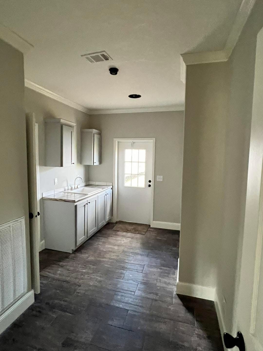 A narrow laundry room with gray walls and dark wood flooring. It features a white countertop, sink, cabinets, and a door with a window, creating a simple, functional space.