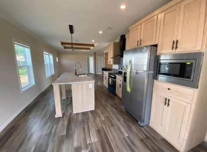Modern kitchen with wood flooring, a central island, and light wood cabinets. Stainless steel fridge and microwave. Bright, spacious, and inviting.