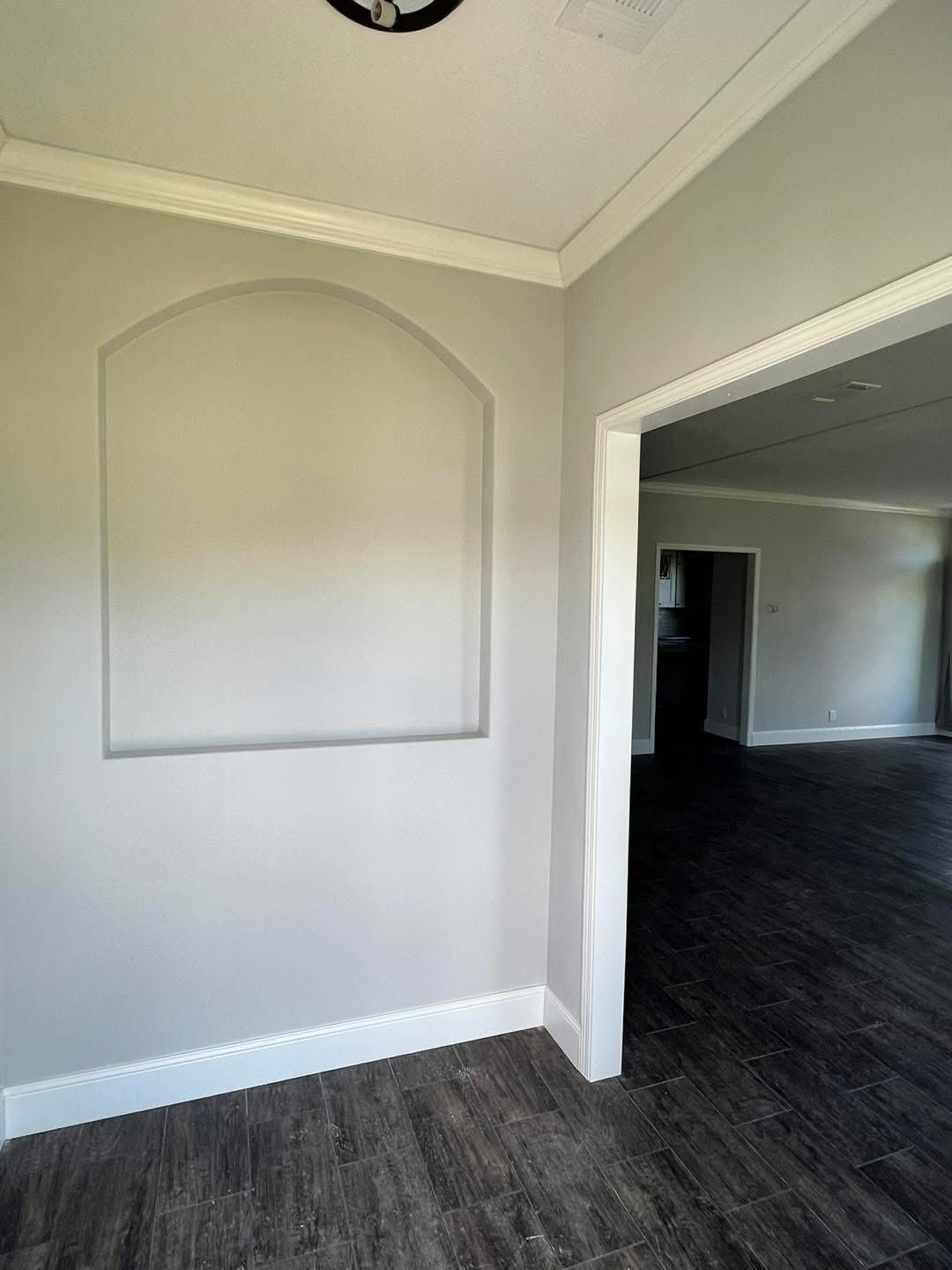 A light gray room with an arch-shaped wall recess, white trim, and dark wood flooring. The space feels modern and minimalist, with an open doorway.