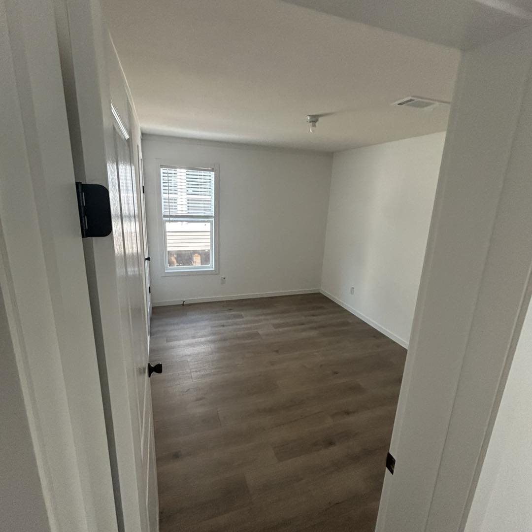 The image shows a small, empty room with white walls and wooden floors. A single window with blinds lets in natural light, creating a calm and minimalist atmosphere.