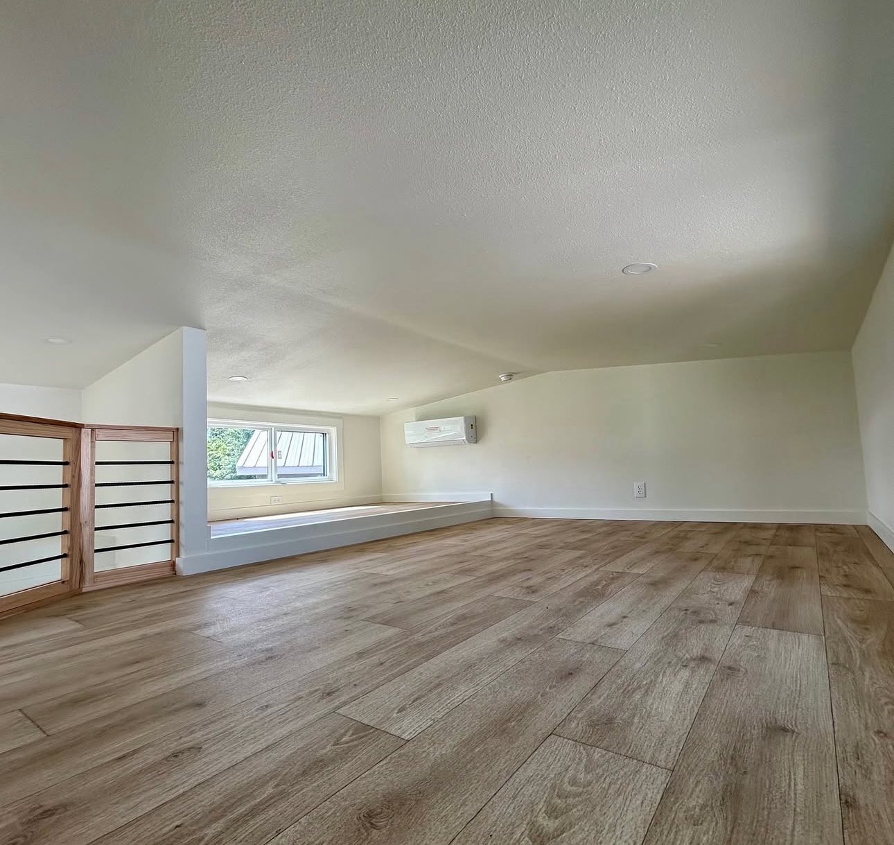 Minimalist attic room with wood flooring, angled white walls, and a small window letting in natural light. An air conditioner is mounted on the wall.