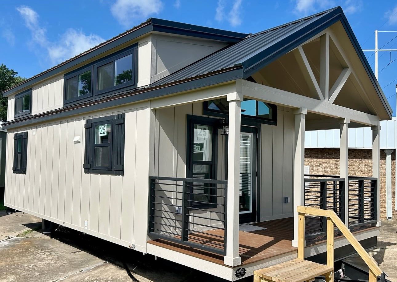 A modern beige tiny house with a black metal roof and loft windows. It has a porch with dark railings and a small wooden staircase, set against a blue sky.