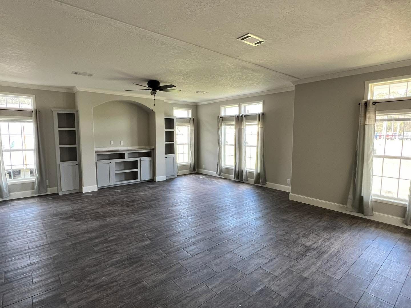 Spacious living room with large windows, gray curtains, and natural light. Features dark wood flooring, built-in shelves, and a ceiling fan. Cozy, inviting ambiance.