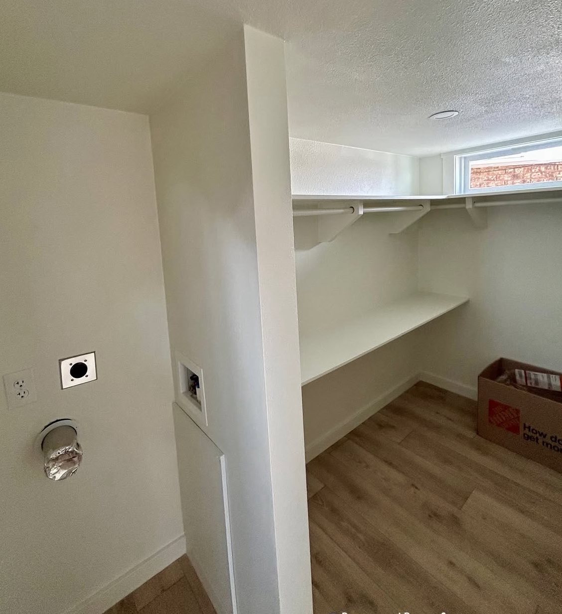 Small laundry room with white walls, featuring a wood floor and utility hookups. A cardboard box sits on the floor under a shelf near a small window.