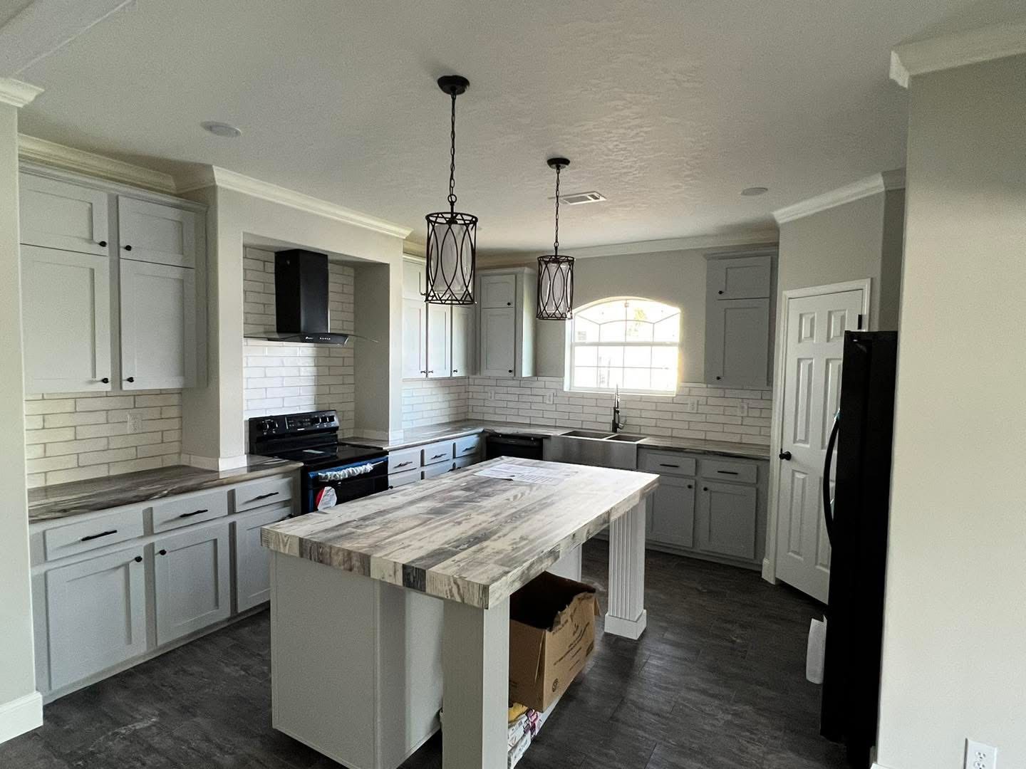 Modern kitchen with light gray cabinets, a large wooden island, and two pendant lights. Brick backsplash, dark stove, and window over the sink.