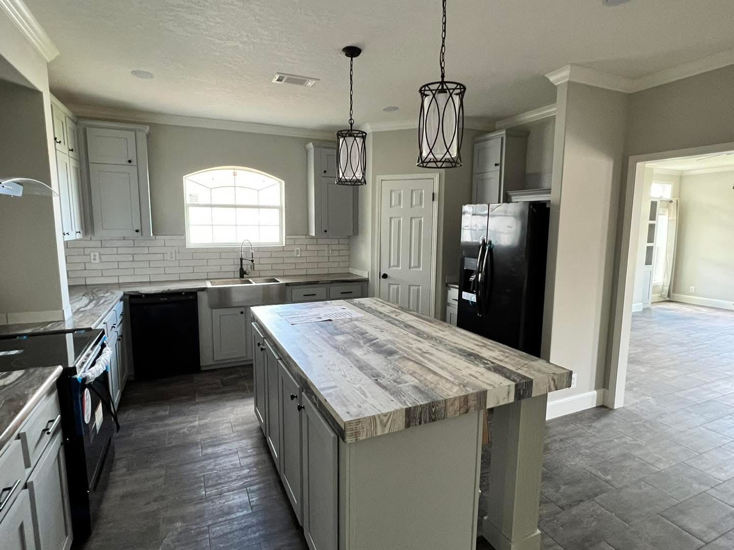 A modern kitchen with light gray cabinets, dark appliances, and a central island with a wood-patterned countertop. Two elegant pendant lights hang above the island, and a large window over the sink provides natural light, creating a bright, inviting atmosphere.