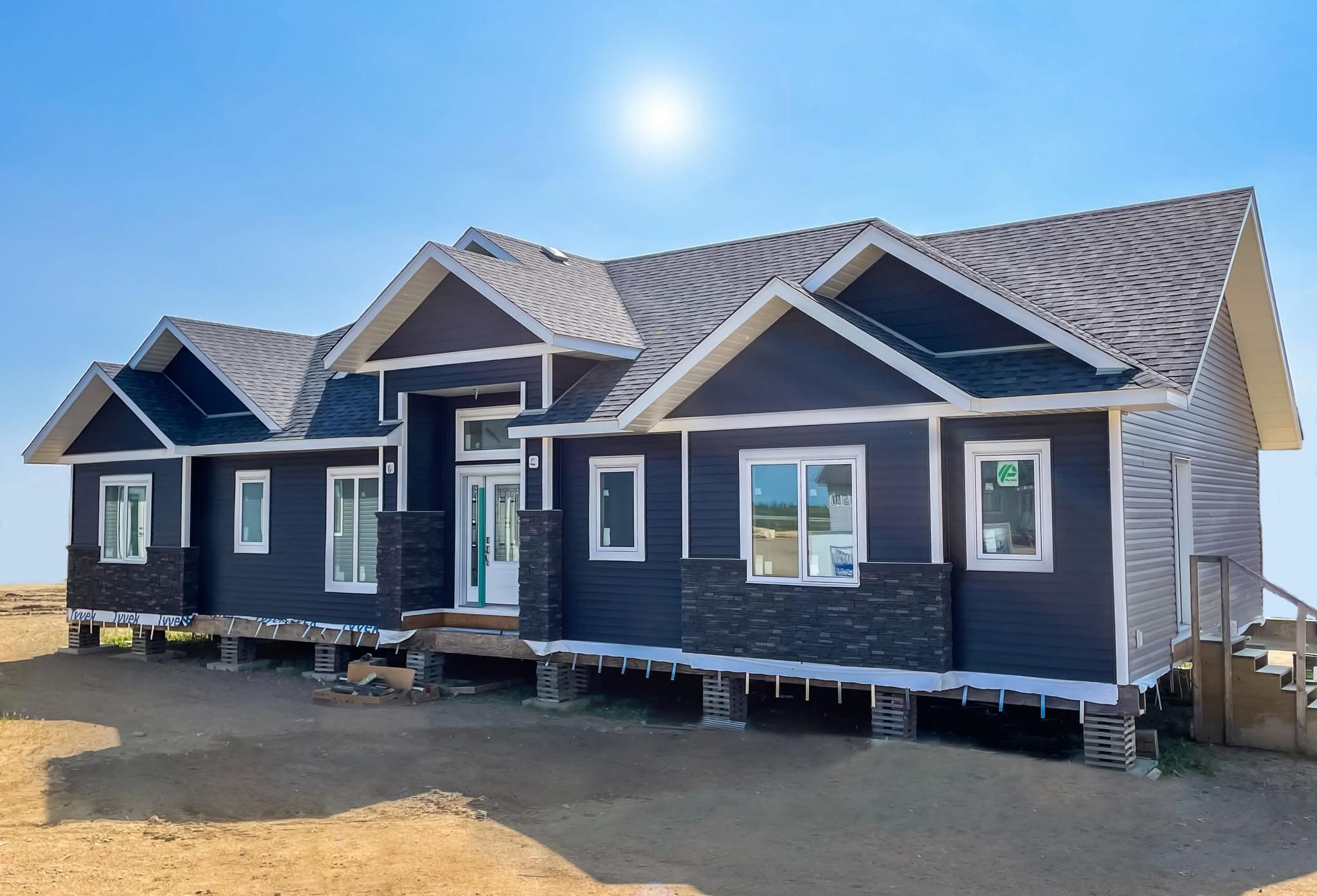 A modern, dark blue, single-story house with a gabled roof stands under a clear blue sky and bright sun, conveying a fresh, welcoming atmosphere.