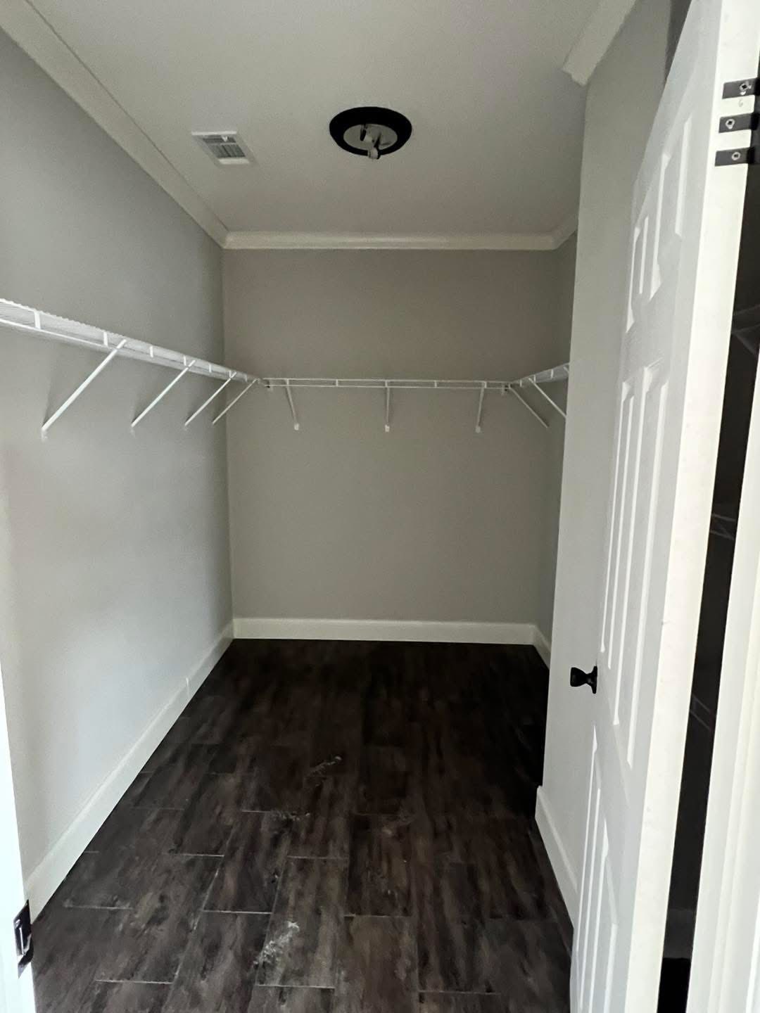 Empty walk-in closet with light gray walls, dark wood flooring, and white wire shelves on three sides. A ceiling light fixture is centered above.