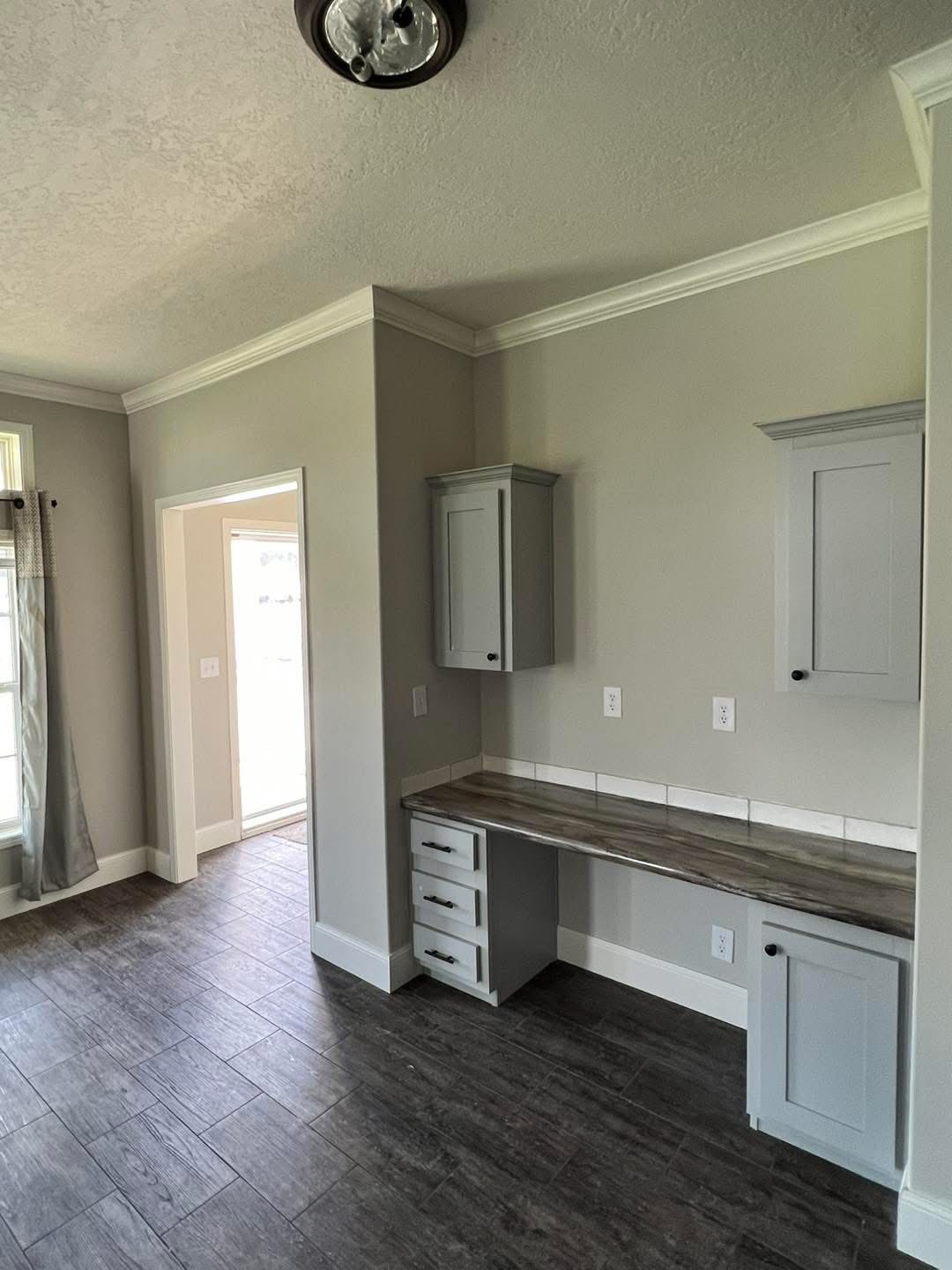 A modern room with gray walls, tile flooring, and minimalist cabinets. A workspace is integrated into one corner, with natural light diffusing in.