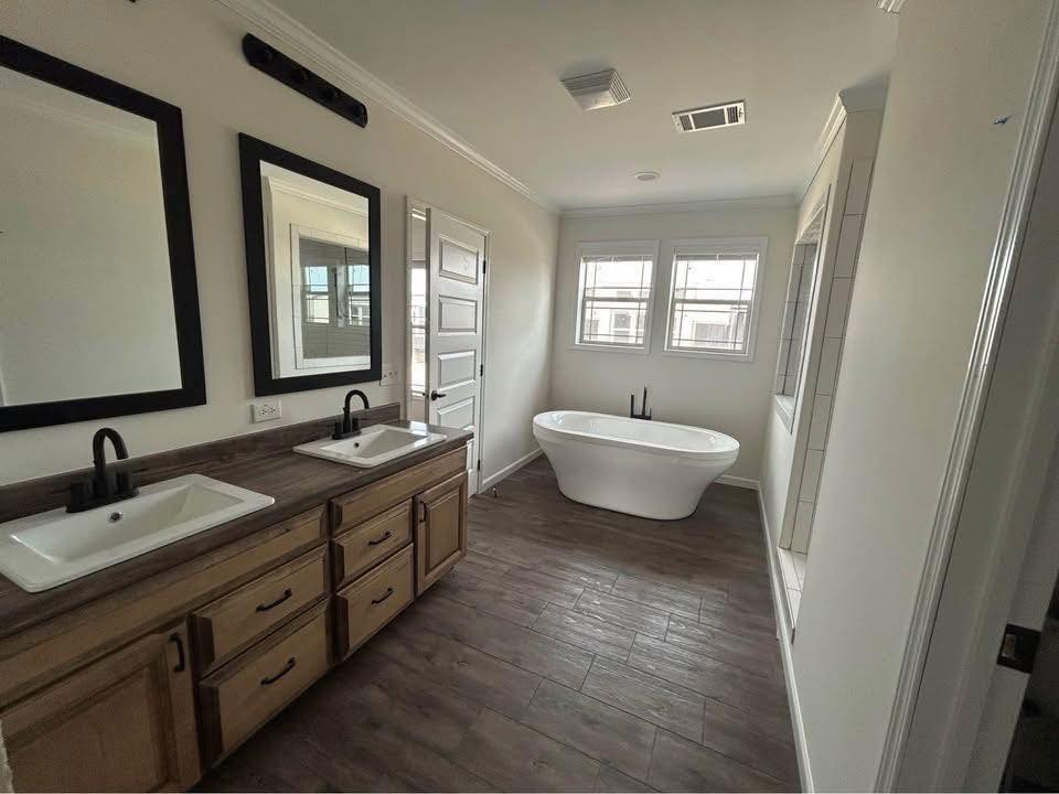 A modern bathroom with dual sinks and black-framed mirrors on the left, a freestanding white bathtub by large windows on the right, and wood-style flooring. Cozy and contemporary.