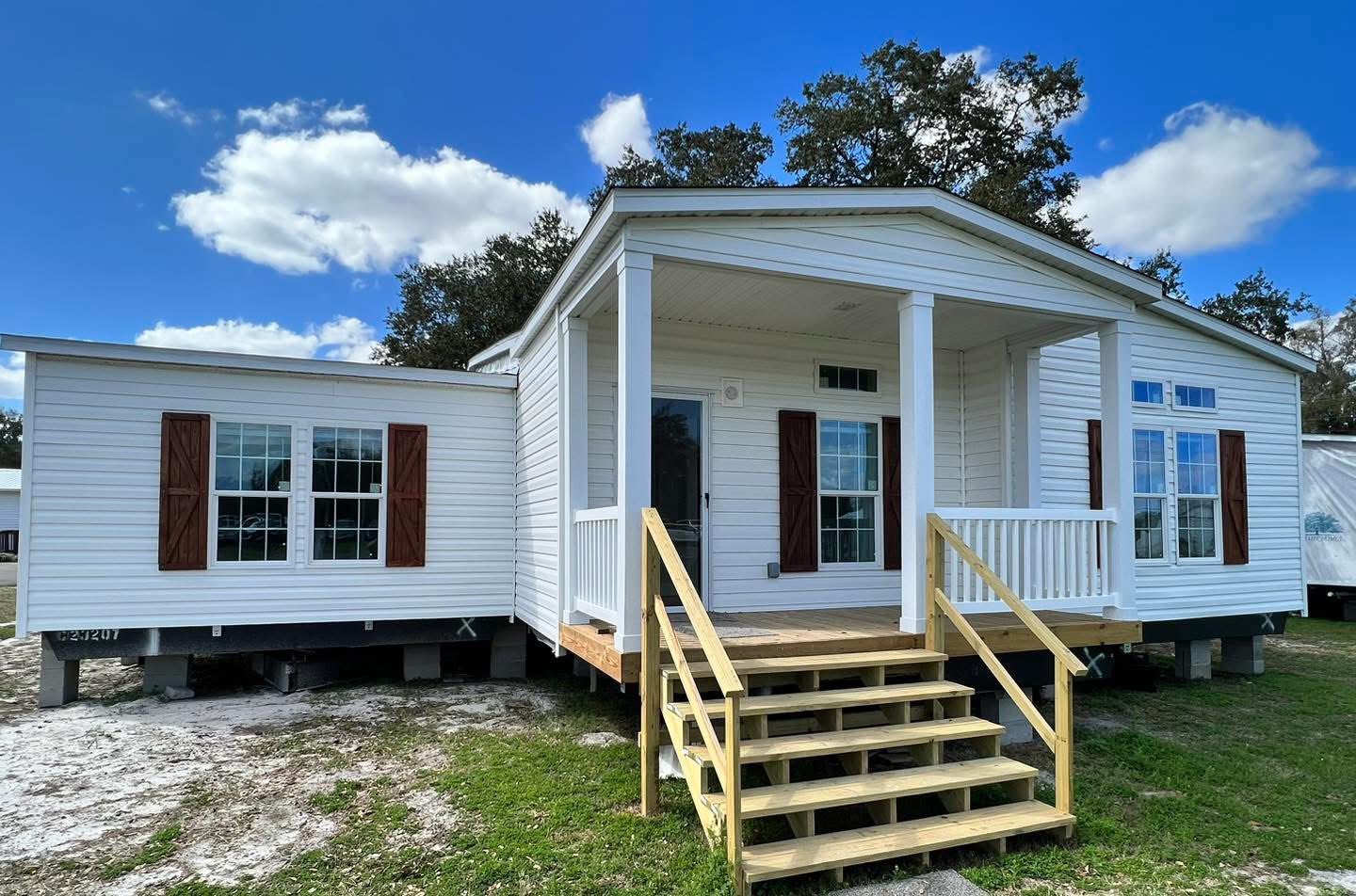 A bright white manufactured home with a small porch and wooden steps. It has several windows with brown shutters, under a clear blue sky and trees.