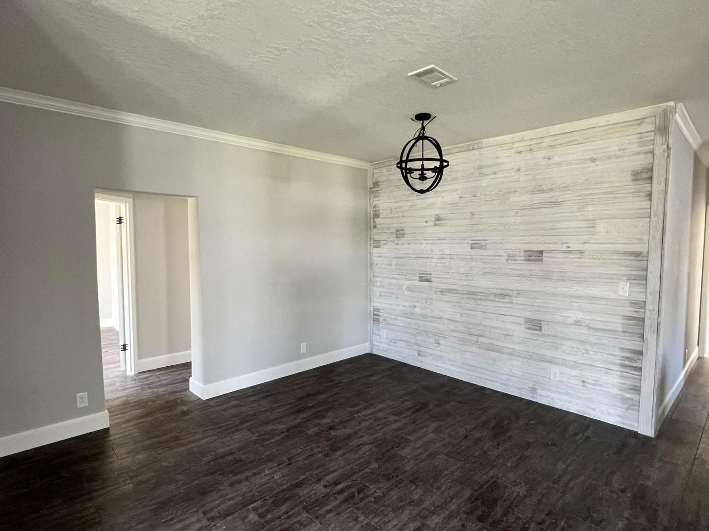 Empty room with dark wood floors, a light gray accent wall, and a modern black orb chandelier. An open doorway leads to another room. The space feels modern and minimal.