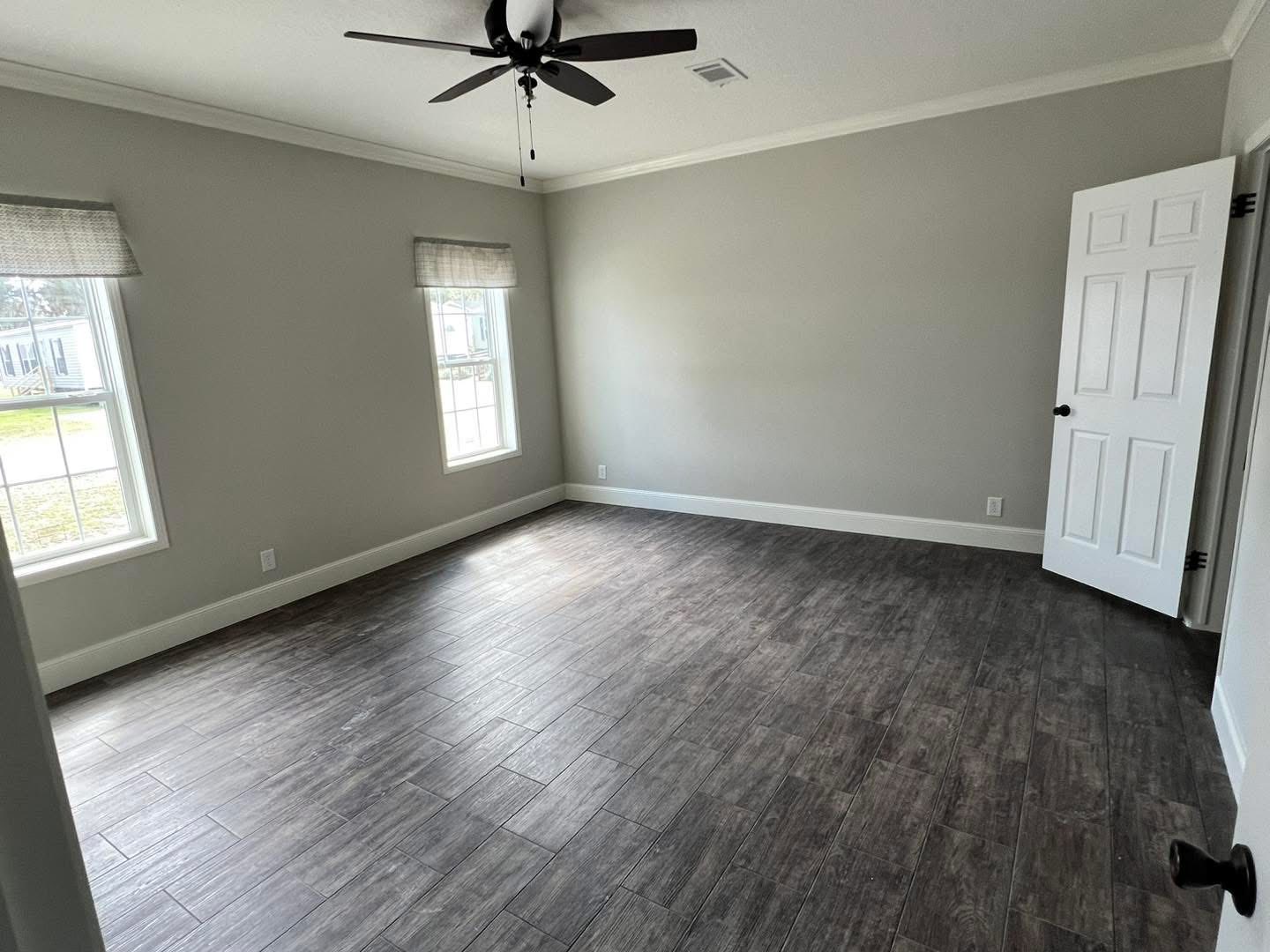 An empty room with two windows, light grey walls, and dark hardwood floors. A white door is slightly open, and a black ceiling fan is installed above.