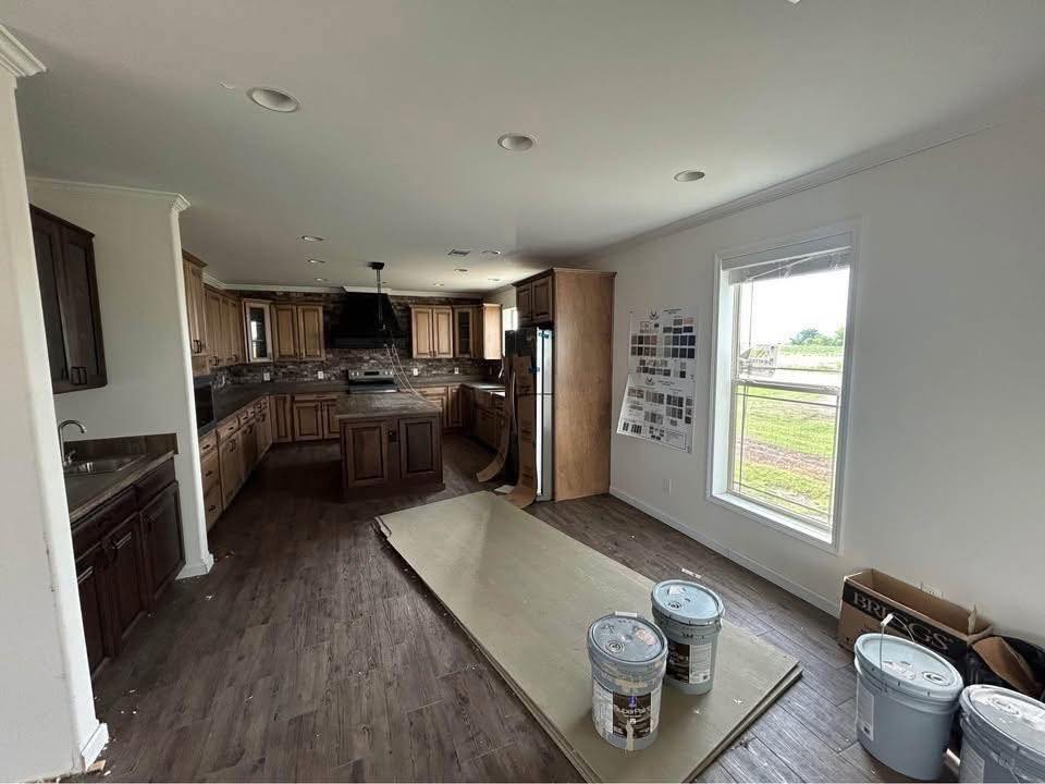 Spacious kitchen interior during renovation with wooden cabinetry and island. Paint cans and boards on hardwood floor, window showing greenery outside.