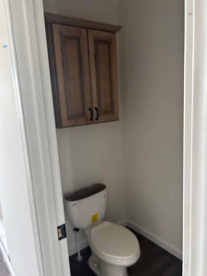 A small bathroom features a white toilet beneath wooden cabinets, set against plain walls and dark flooring. The scene is minimalistic and functional.