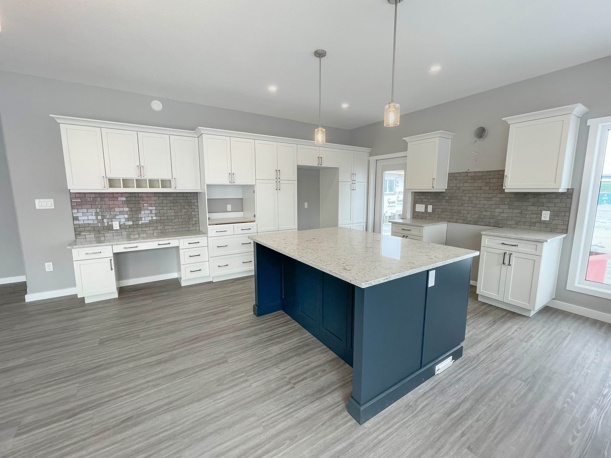Modern kitchen with white cabinets, gray tiled backsplash, and a large island with a dark base and light countertop. Pendant lights hang above.