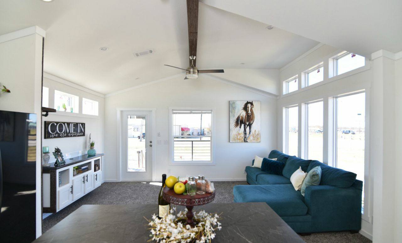 A modern living room with vaulted ceilings and a ceiling fan. Large windows illuminate a teal sectional sofa and horse artwork. A sign reads "Come in, you are awesome."