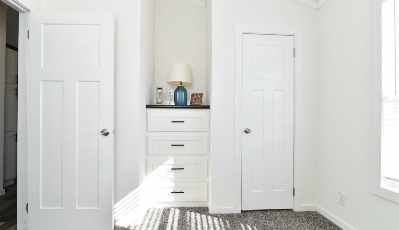 Bright, minimalist room with white walls and doors. Features built-in drawers and decor like a blue vase and lamp; sunlight streams in, casting shadows.