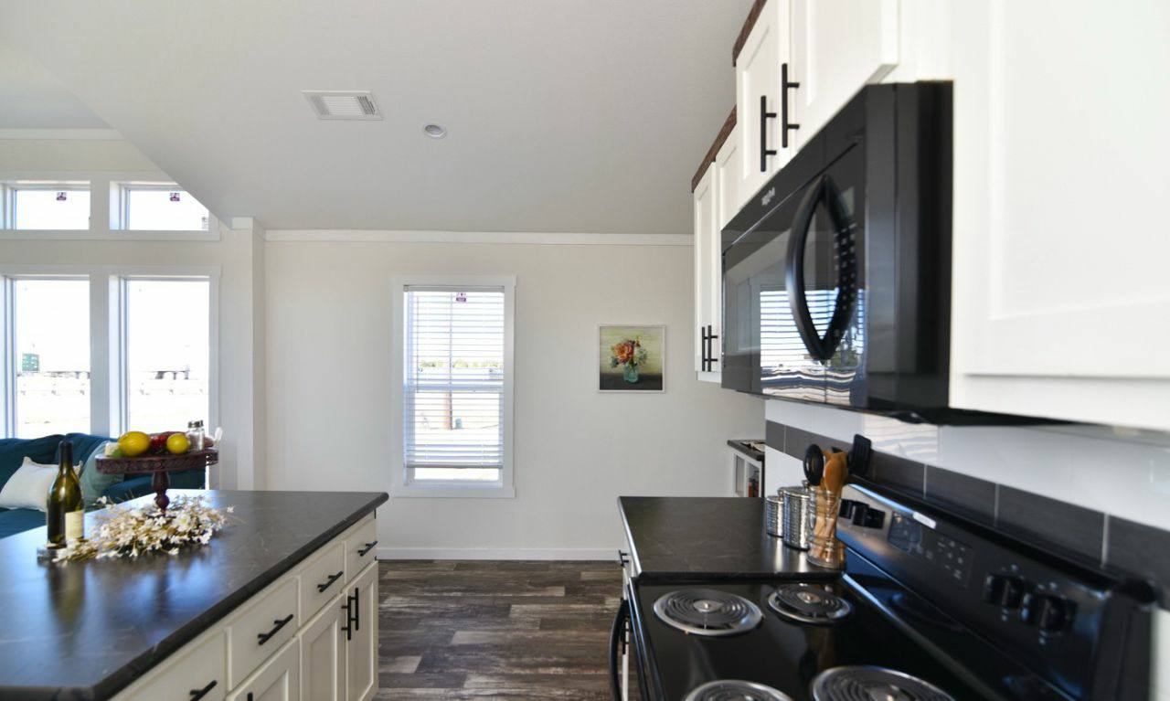 Modern kitchen with white cabinets, black countertops, and appliances. Sunlight streams through large windows, creating a bright, inviting atmosphere.