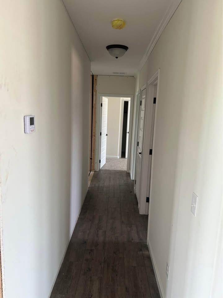 Narrow hallway with wooden floor, white walls, and two ceiling lights. Several closed doors on the right lead to carpeted areas, creating a simple, clean ambiance.