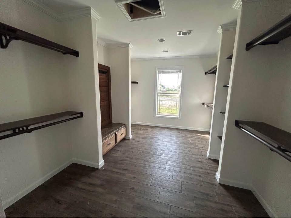 Spacious walk-in closet with wooden shelves and drawers against off-white walls. A large window allows natural light, highlighting the dark wood floor.