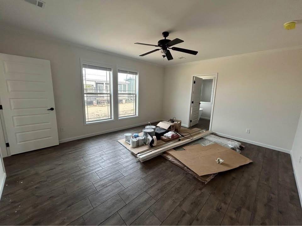 Unfurnished room with wood flooring, white walls, and a ceiling fan. Materials like paint cans and wood planks are piled on cardboard in the center. Large windows and an open door lead to another room, suggesting renovation.