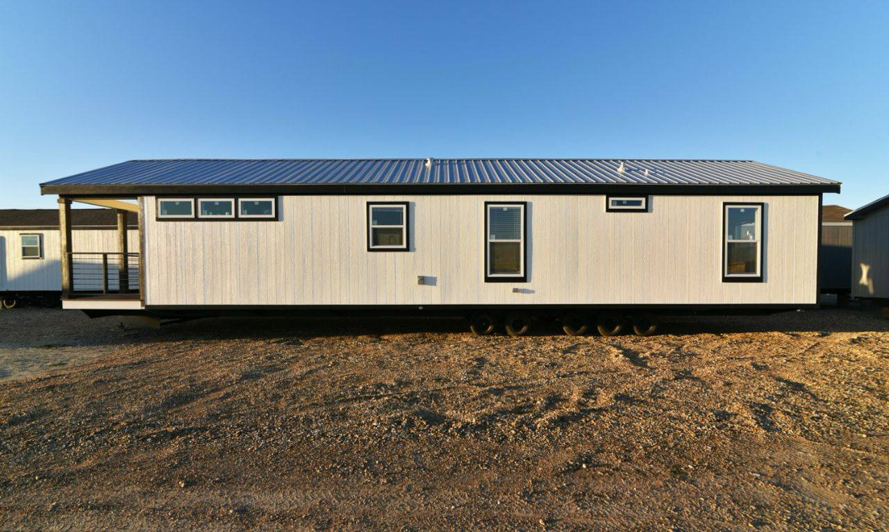 A modern mobile home with white siding and a sloped metal roof, set on a gravel lot. Large windows are evenly spaced, creating a clean, minimalist look.