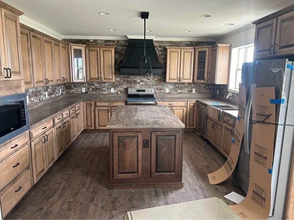 Spacious kitchen with wood cabinets, a central island, and brick backsplash. Stainless steel appliances partially unpacked, creating a fresh, rustic tone.