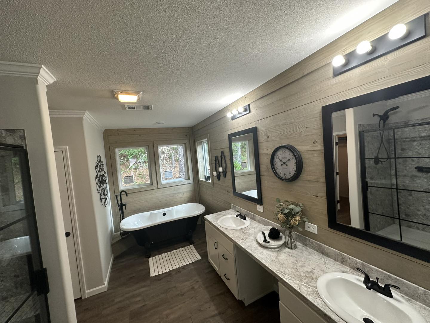 A modern bathroom features a freestanding black tub by large windows, dual sinks with a marble countertop, wooden walls with mirrors, and ambient lighting.