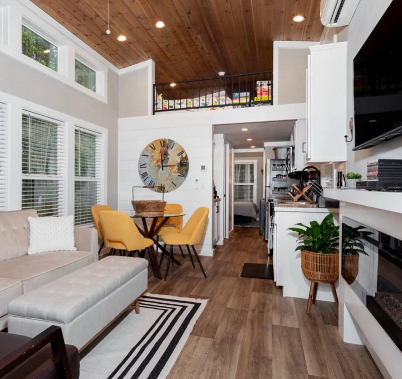 Modern tiny house interior with wooden ceiling, featuring a beige sofa, yellow chairs around a circular table, and a large clock. Cozy ambiance.