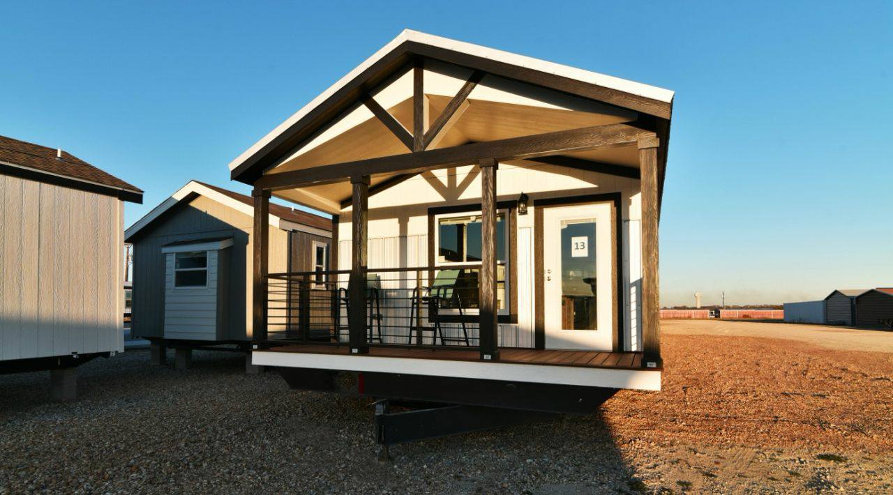Small, modern tiny house with a covered porch and dark wood beams, set on a gravel lot under a clear blue sky, conveying simplicity and warmth.