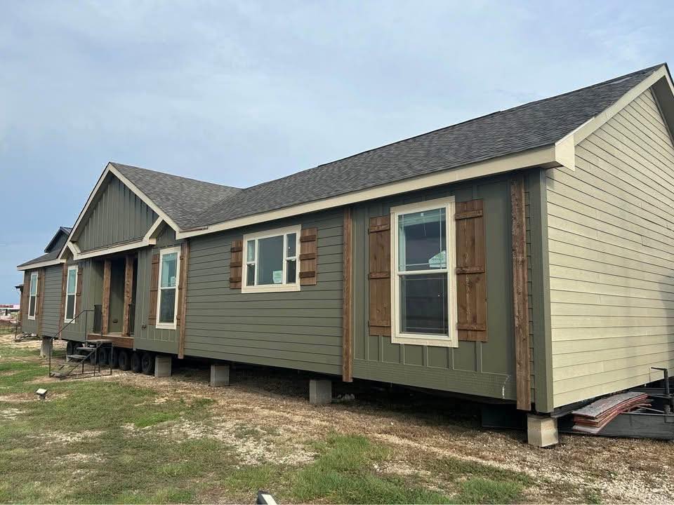 Long, single-story manufactured home on a grassy lot under a clear sky. It has green siding, wooden shutters, and a dark shingled roof, conveying simplicity.