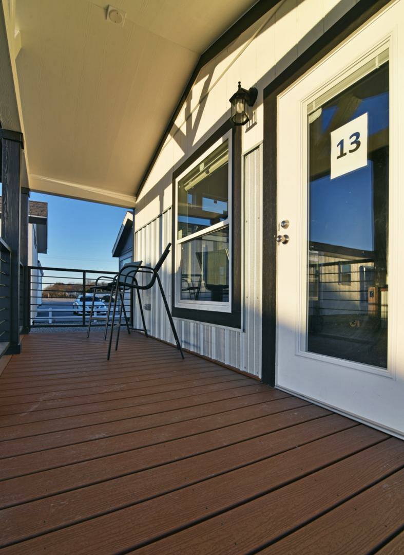 Covered porch with wooden floorboards features a metal railing, two high chairs, and a door labeled 13. Bright sunlight casts shadows, evoking a peaceful atmosphere.